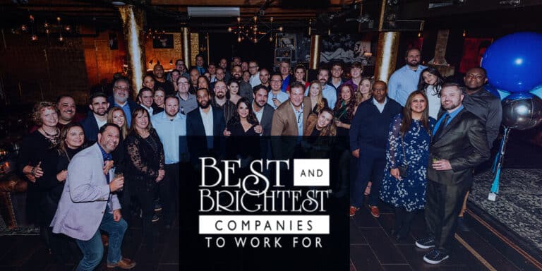 A large group of professionally dressed people pose for a photo at an indoor event. In front, a sign reads "Best and Brightest Companies to Work For." Blue balloons are visible on the right side.