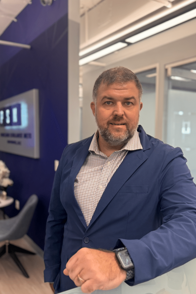 A man with short gray hair and a beard, wearing a blue blazer over a checked shirt and a smartwatch, stands indoors near a modern office area with glass walls and a sign in the background.