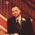 A man with short hair and a beard, wearing a black tuxedo with a white rose boutonniere, sits in front of an elegant staircase with ornate railing and red walls.