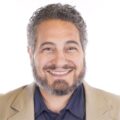 A man with curly salt-and-pepper hair and a beard smiles at the camera. He is wearing a tan blazer over a dark shirt and is pictured against a white background.