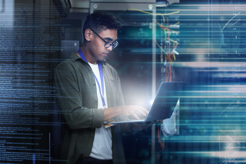 A person wearing glasses stands in a server room typing on a laptop, surrounded by digital code and blue light effects, suggesting cybersecurity or information technology work.