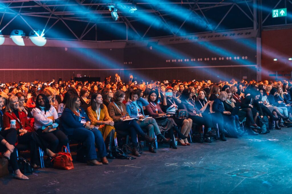 A large audience sits in rows at an indoor event or conference, illuminated by blue stage lights. Most attendees appear to be women, and many are engaged, watching the stage or clapping.
