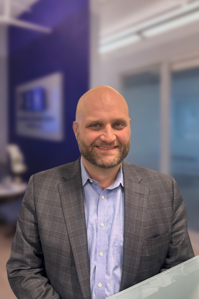 A smiling bald man with a beard, wearing a blue shirt and gray plaid blazer, stands in a modern office with blue and white walls and glass-paneled rooms in the background.