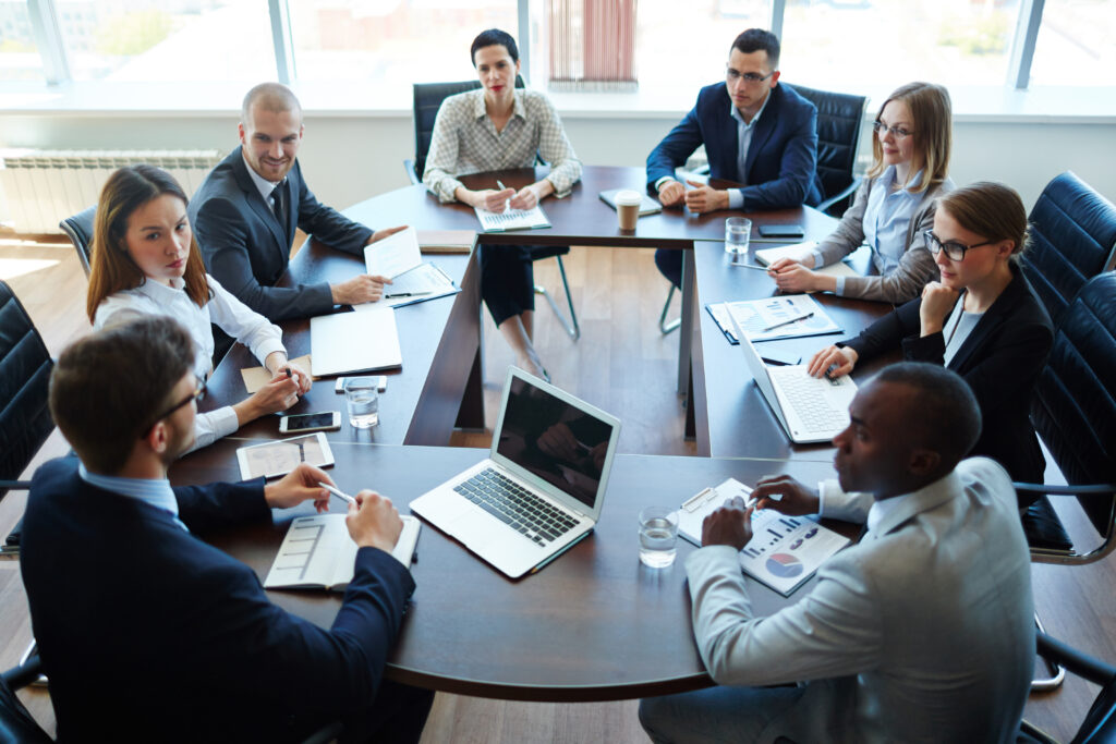 Eight people in business attire sit around a conference table in a modern office, engaged in a meeting with laptops, documents, and charts visible on the table. Large windows let in natural light.