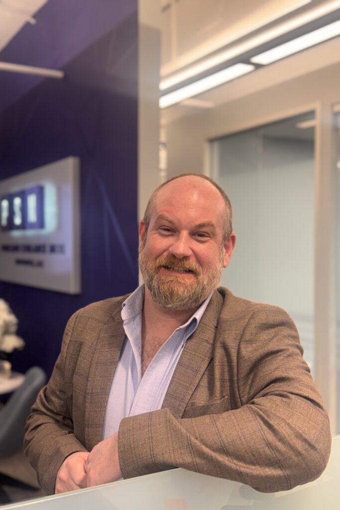 A bearded man in a brown blazer and light blue shirt smiles while leaning on a glass counter in a modern office with a sign and seating area in the background.