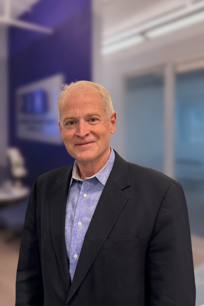 A middle-aged man with short gray hair wears a dark blazer over a light blue shirt, standing in a modern office with glass walls and a blurred background.