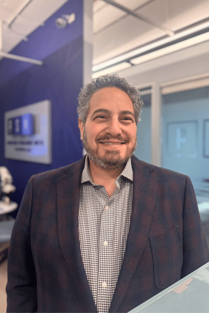 A man with curly hair and a beard smiles while standing indoors, wearing a checked shirt and dark blazer. The background shows a modern office with glass walls and a blue accent wall with an office sign.