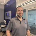 A man with short dark hair and a trimmed beard smiles while standing in an office. He wears a gray polo shirt with black trim and a logo on the chest. The background shows modern office furniture and a wall with framed pictures.