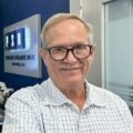 Smiling older man with glasses and a mustache wearing a checked shirt, standing indoors in front of a blue wall with a PSN sign and glass office partitions in the background.