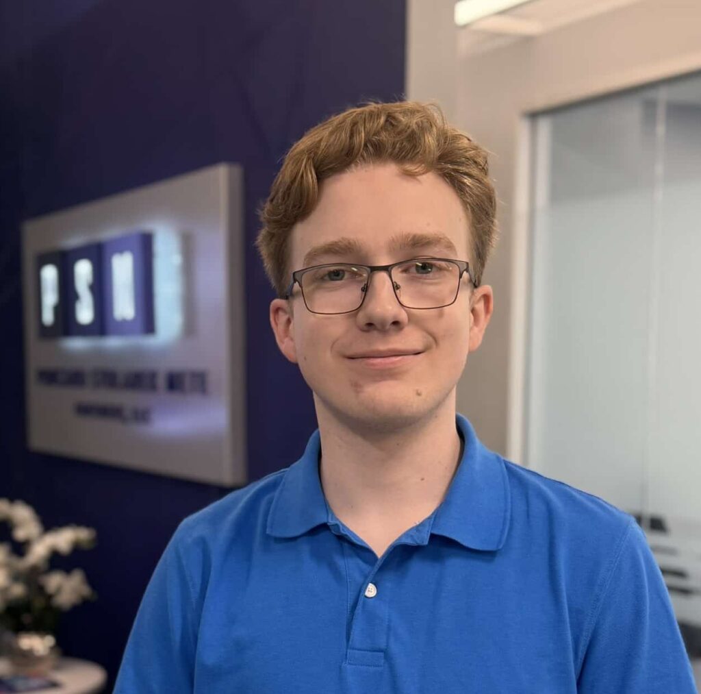 A young person with glasses and red hair, wearing a blue polo shirt, stands in a modern office with a sign reading “PSH” in the background and flowers on a small table.