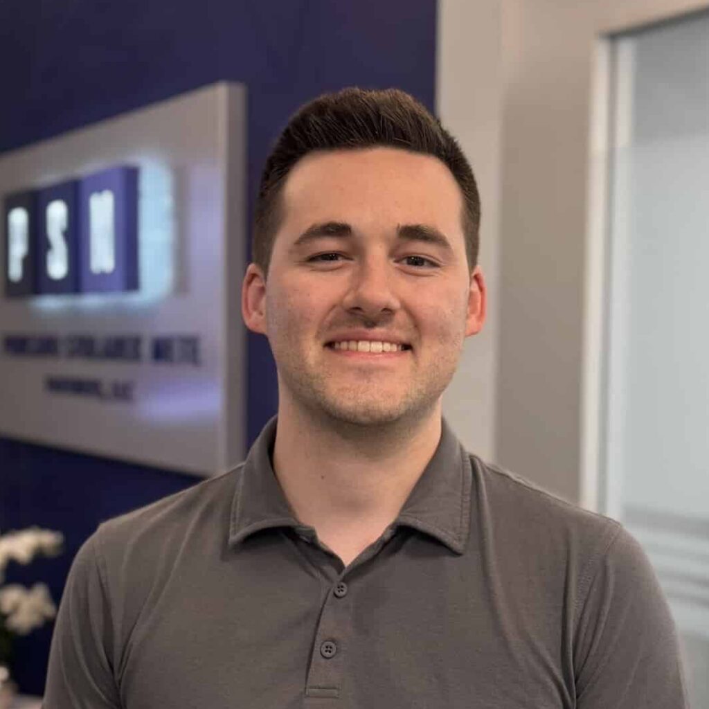 A person in a gray polo shirt stands smiling in a modern office with blue walls, a blurred sign, chairs, flowers, and glass panels in the background.