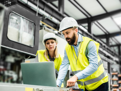 Two workers in safety vests and helmets stand in a factory, looking at a laptop on a worktable. The man is typing while the woman observes. Industrial equipment is visible in the background.